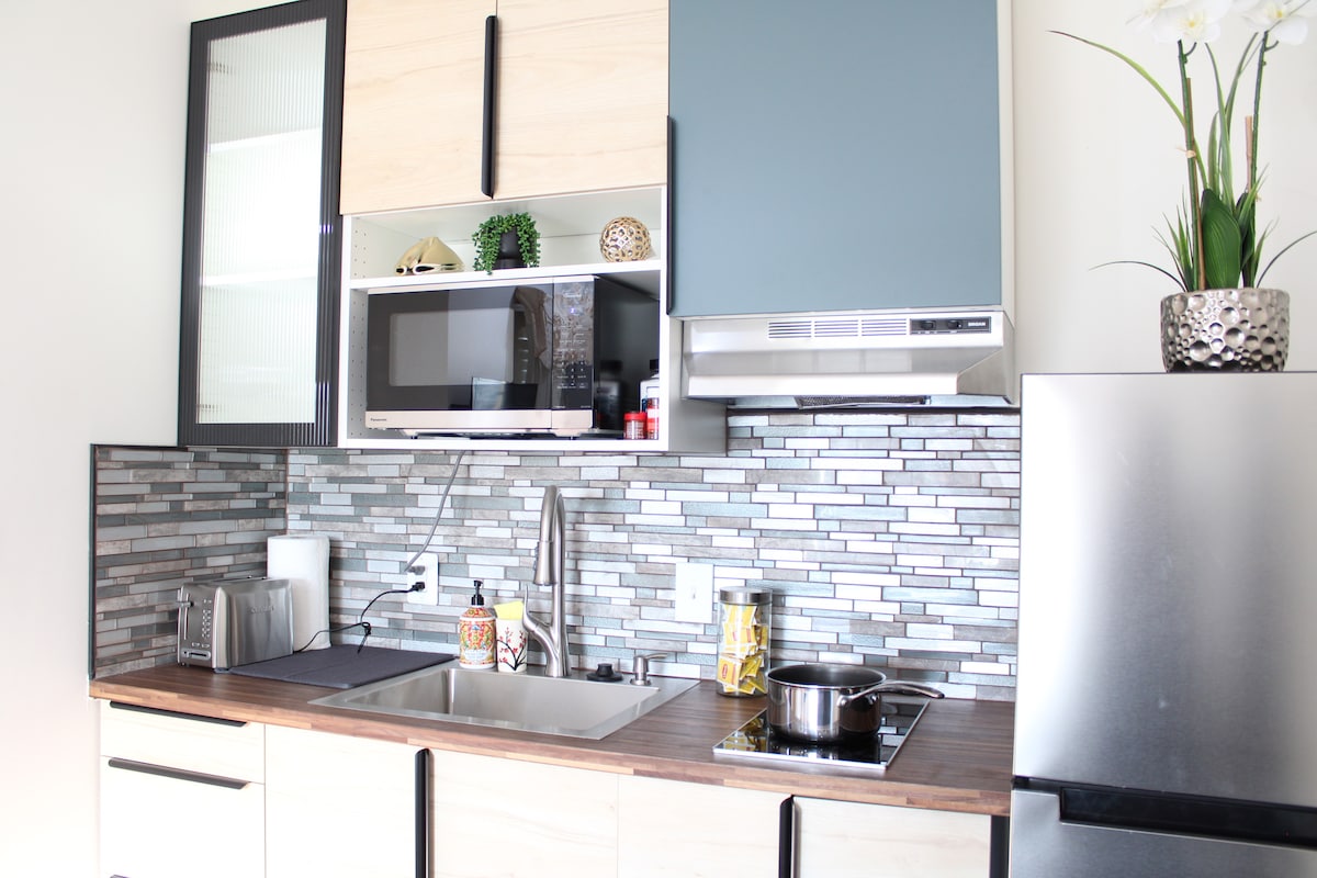 A modern kitchenette is displayed, featuring a stainless steel refrigerator and a sleek microwave. The stylish backsplash consists of mosaic tiles in shades of gray and blue. A stainless steel sink and stovetop are positioned on a wooden countertop, complemented by decorative plants on the shelves.