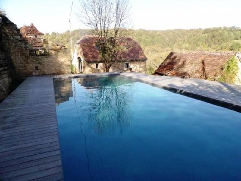 A tranquil swimming pool is nestled among lush greenery, reflecting the surrounding landscape. Wooden decking frames the pool area, enhancing the connection to nature. Stone walls and rustic buildings are visible in the background, contributing to the serene atmosphere.