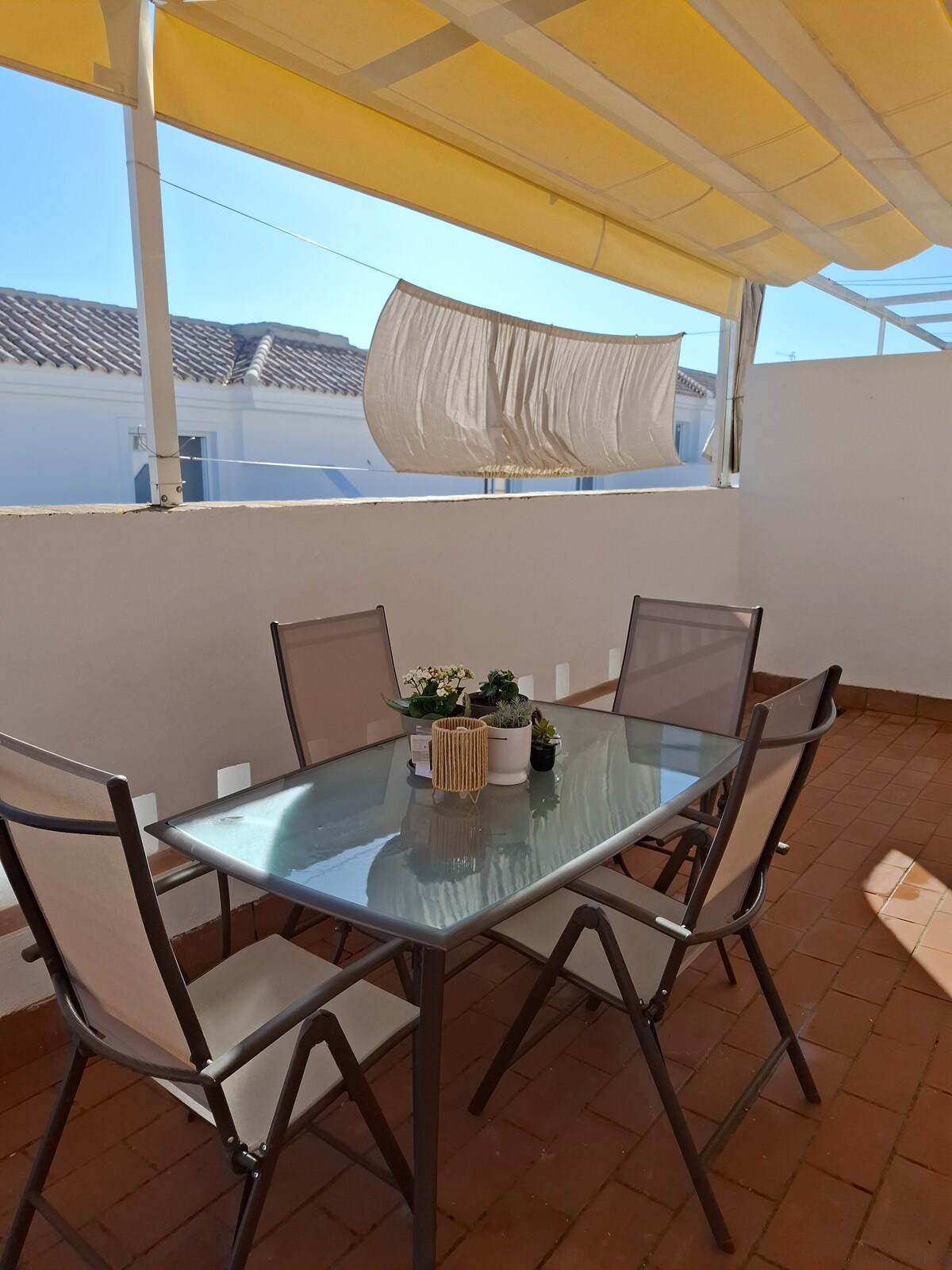 An outdoor terrace space features a glass-top dining table surrounded by four light-colored chairs. A subtle yellow awning provides shade, while potted plants and a decorative item are placed on the table. Bright sunlight illuminates the area, creating a relaxed atmosphere.