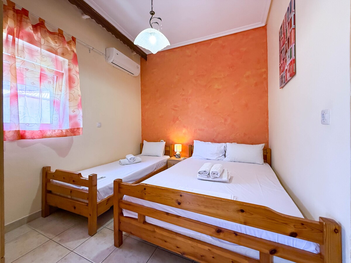 A simply furnished bedroom is presented with a single bed and a double bed, both with white linens. Light walls are complemented by a vibrant orange accent wall. A small lamp is positioned on a bedside table, and a window with sheer curtains allows natural light to enter the space.