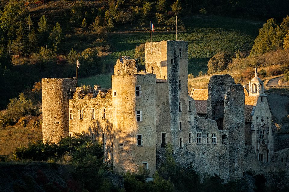 An ancient stone castle is nestled among a hilly landscape, partially illuminated by the setting sun. The structure showcases weathered walls, several turrets, and flags waving atop. Verdant hills surround the castle, creating a scenic backdrop that enhances the historic architecture.