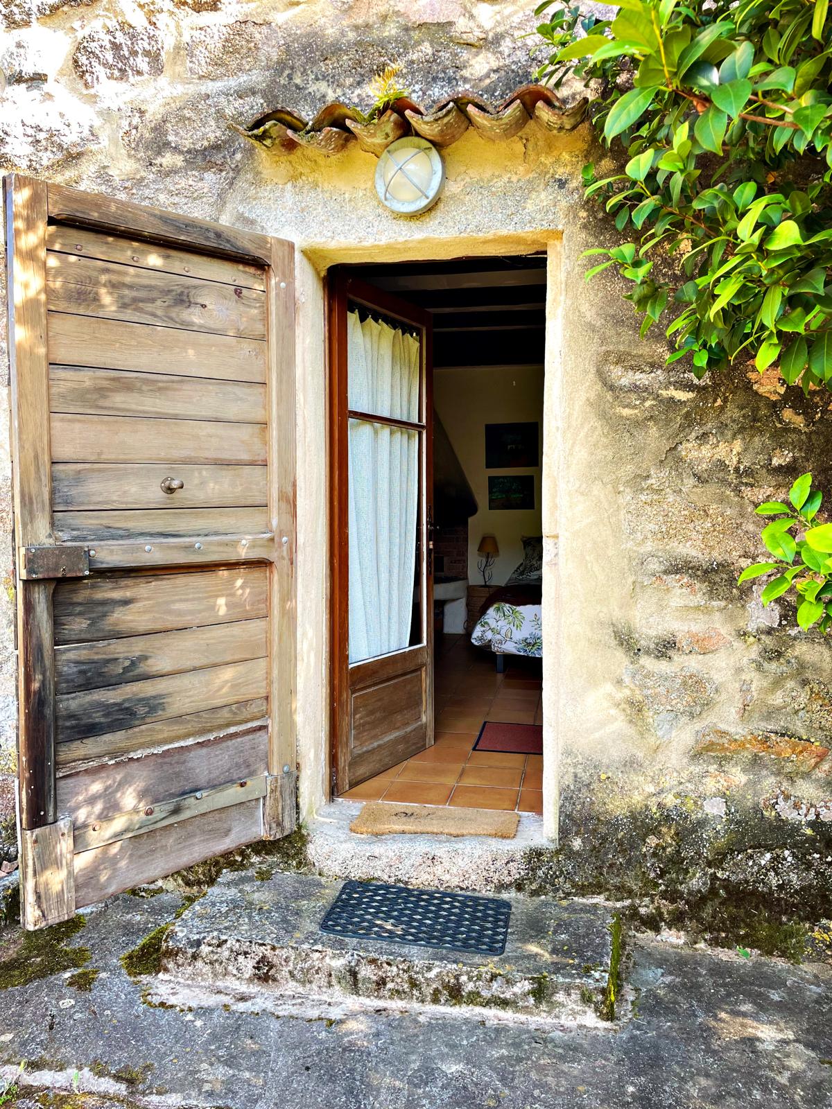 The entrance showcases a rustic wooden door, partially opened, revealing a glimpse of the inviting interior. A small mat is placed at the threshold, and sunlight filters through the white curtains, highlighting the stone wall and the surrounding greenery.