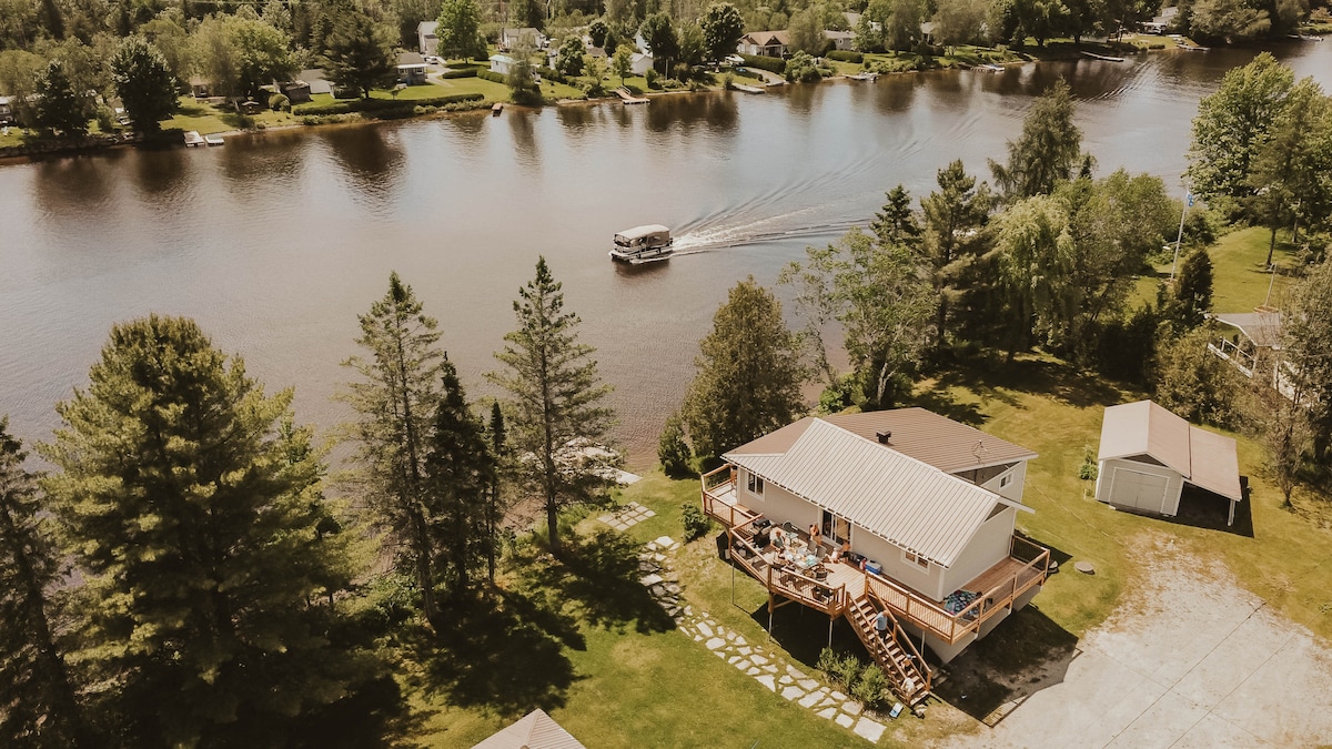 The chalet is situated on a spacious grassy area adjacent to the Saint-François River. A wooden deck extends from the home, offering seating for outdoor enjoyment. A boat can be seen gliding along the calm waters, surrounded by trees and well-maintained properties in the background.