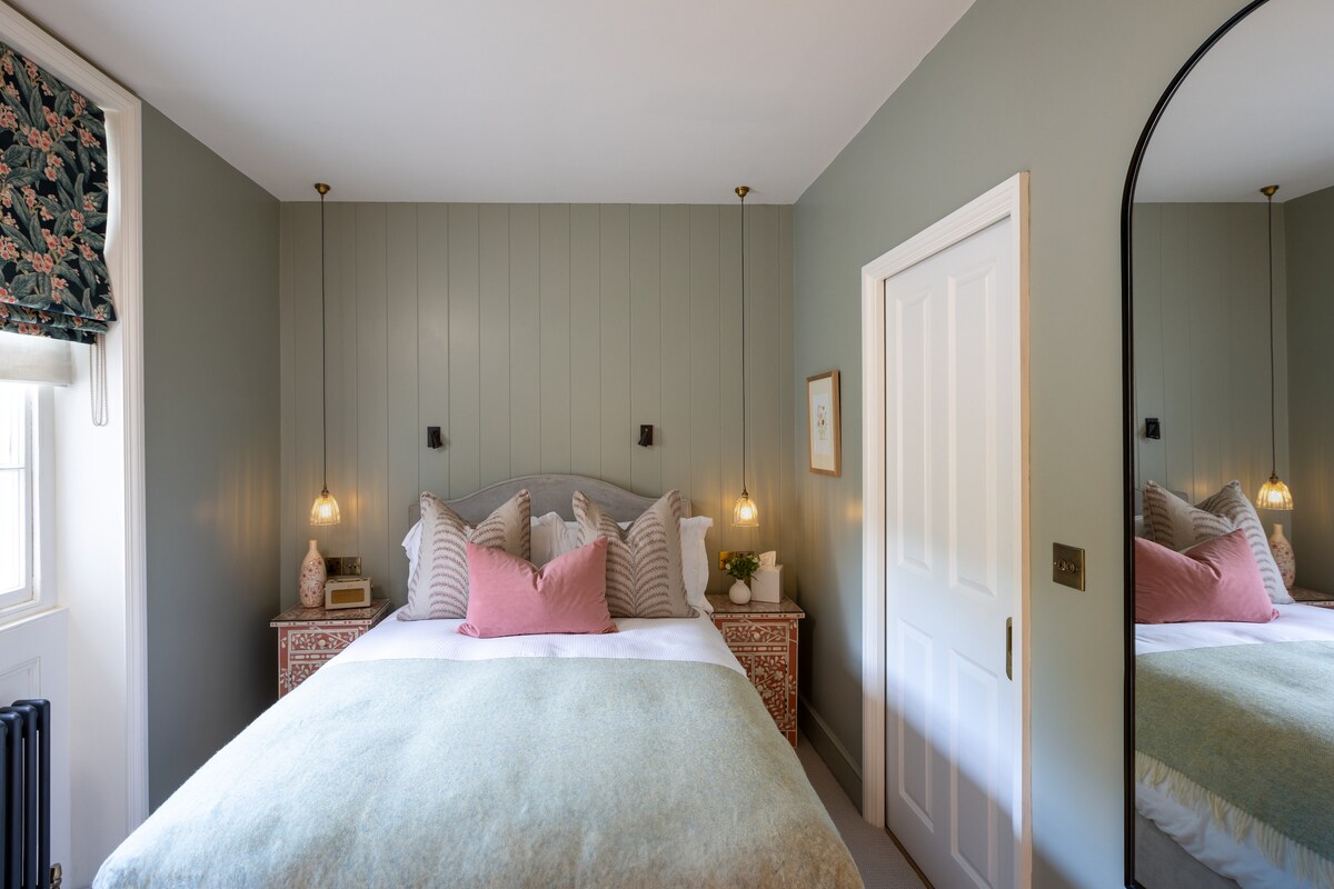 The bedroom features a spacious bed adorned with decorative pillows and a light blanket. Stylish bedside lamps provide soft lighting, and an ornate mirror enhances the room’s charm. Neutral wall colors and a patterned window treatment lend a cozy feel to the inviting space.