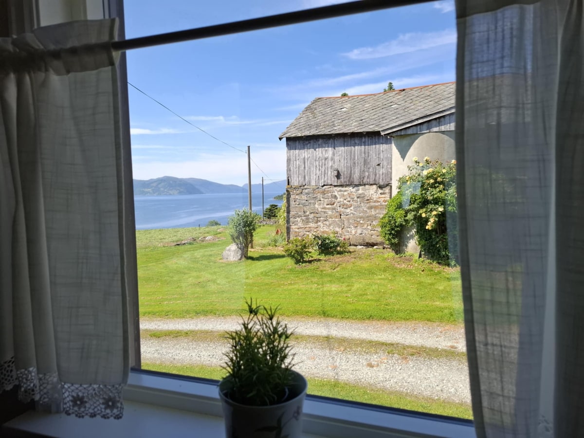 A view from the window showcases a green lawn leading to a stone building and the serene water of Nordfjorden. The gentle curves of the landscape are framed by sheer curtains, while the sky above is clear with soft clouds.