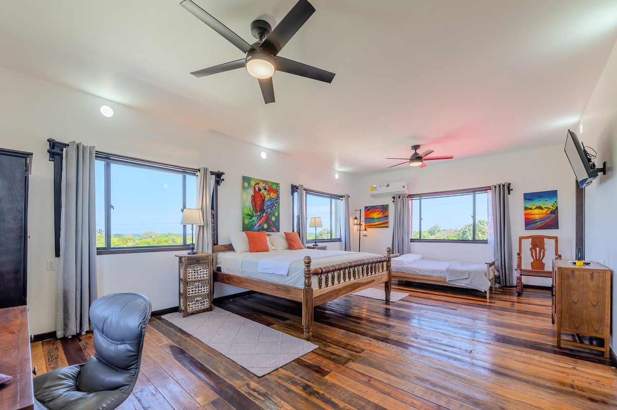 A spacious bedroom features large windows showcasing distant mountain and sea views. Two beds are positioned within the room, with one prominently displayed on a wooden frame. Light wooden flooring and colorful wall art complement the bright, airy atmosphere. A desk and a flat-screen TV are also present.