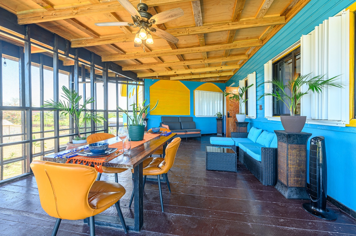 A spacious porch area is featured, with a large wooden dining table surrounded by colorful chairs. Comfortable seating is arranged with cushions, complemented by tropical plants. The space is brightened by natural light streaming through large windows, enhancing the inviting atmosphere.