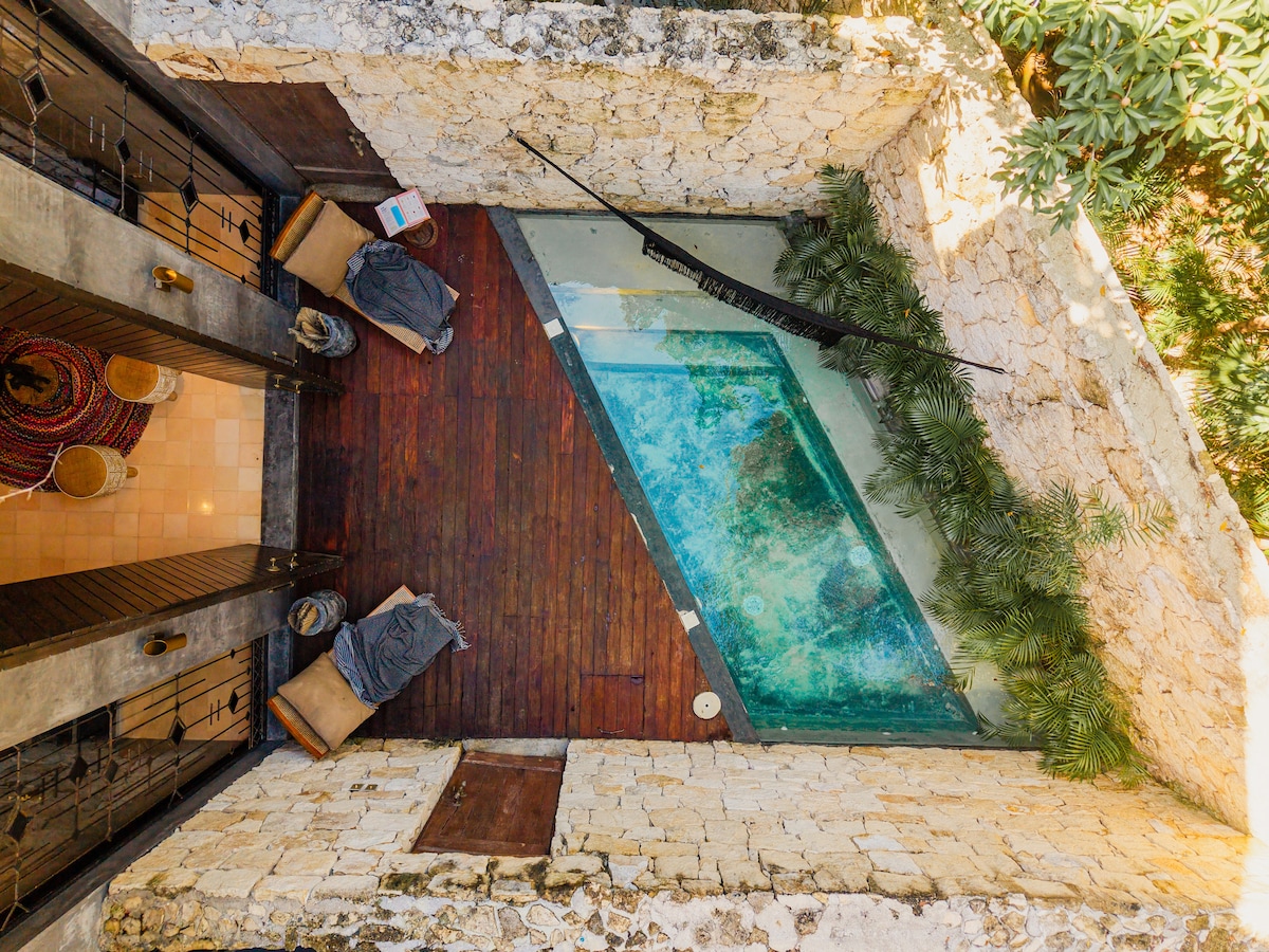 An aerial view showcases a private pool framed by lush greenery and wooden decking. Hammocks are draped between the walls, offering a tranquil spot for relaxation. The natural stone walls and wooden accents provide a rustic feel, enhancing the outdoor oasis.