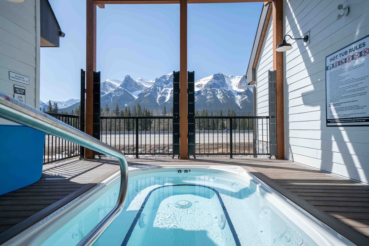 The image showcases an outdoor communal hot tub framed by a wooden structure. Snow-capped mountains are visible in the background, enhancing the natural beauty of the setting. Clear blue skies and wooden decking create a serene environment for relaxation.