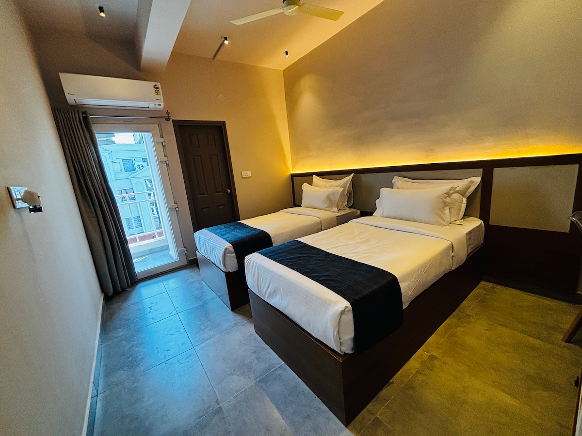 The room features two cozy twin beds with comfortable bedding, positioned against a stylish backdrop. A large window allows natural light to fill the space, leading to a private balcony. Soft lighting enhances the modern design, creating a serene environment for relaxation.