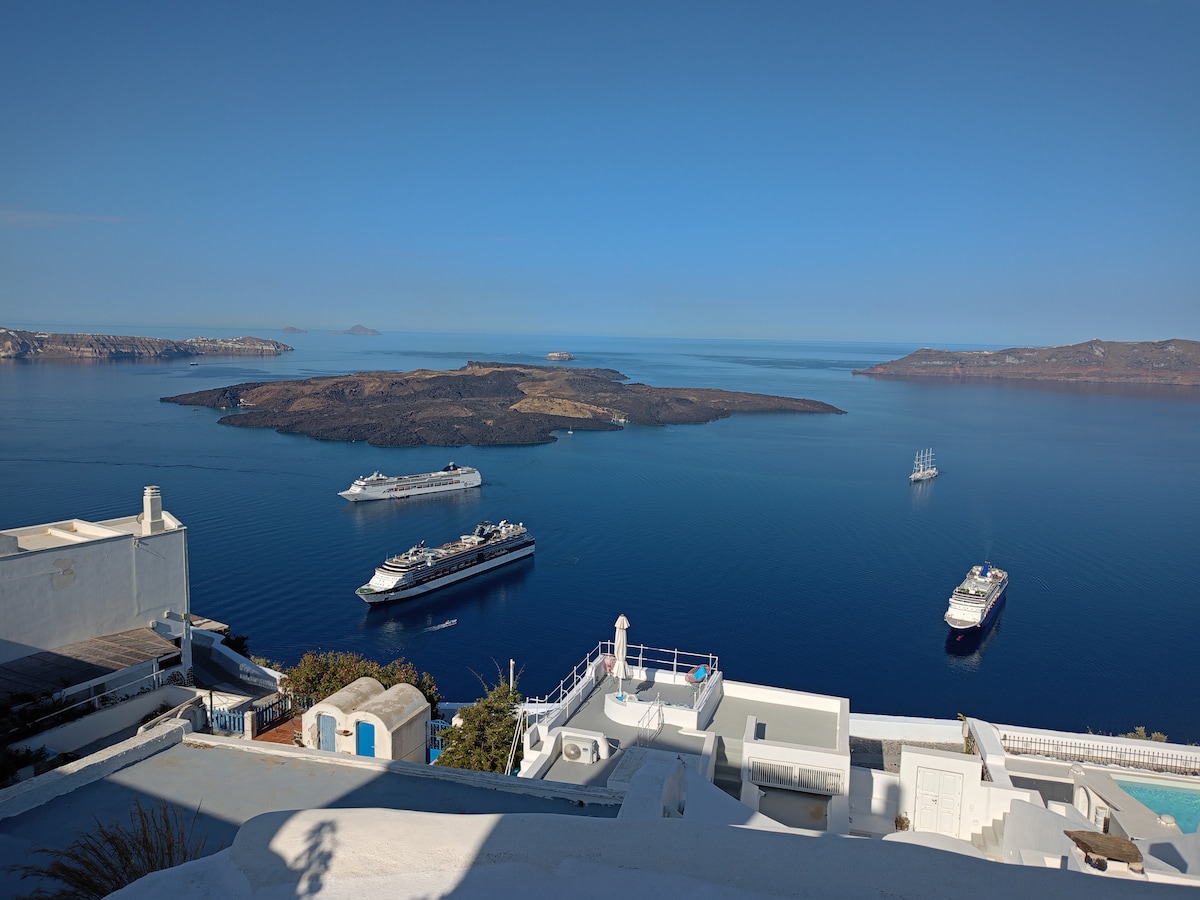 A panoramic view captures the serene caldera, featuring calm blue waters with several boats anchored. Small islands dot the horizon against a clear sky, while white structures of nearby homes blend harmoniously with the landscape, emphasizing the peaceful surroundings of Firostefani.