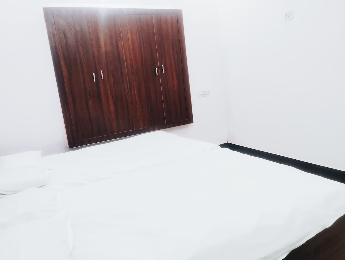 A simple bedroom features a spacious bed covered with white bedding. Wooden wardrobe doors are mounted on the wall, providing storage. The room is primarily adorned in soft, light colors, promoting a clean and peaceful environment.
