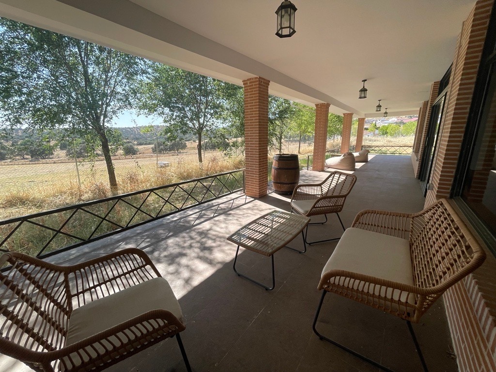 A spacious porch is lined with several wicker chairs and small tables, offering a place to relax. Natural light filters through the trees outside, enhancing the connection to the surrounding landscape.