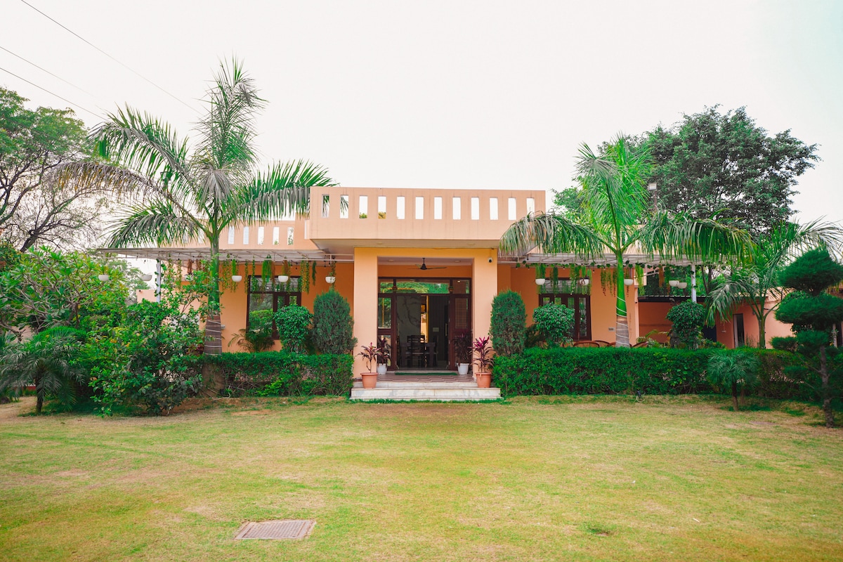A serene farmhouse exterior is framed by lush greenery and palm trees. The structure features a wide entryway with a set of steps leading up to a covered porch. A manicured lawn stretches out in front, enhancing the inviting atmosphere.