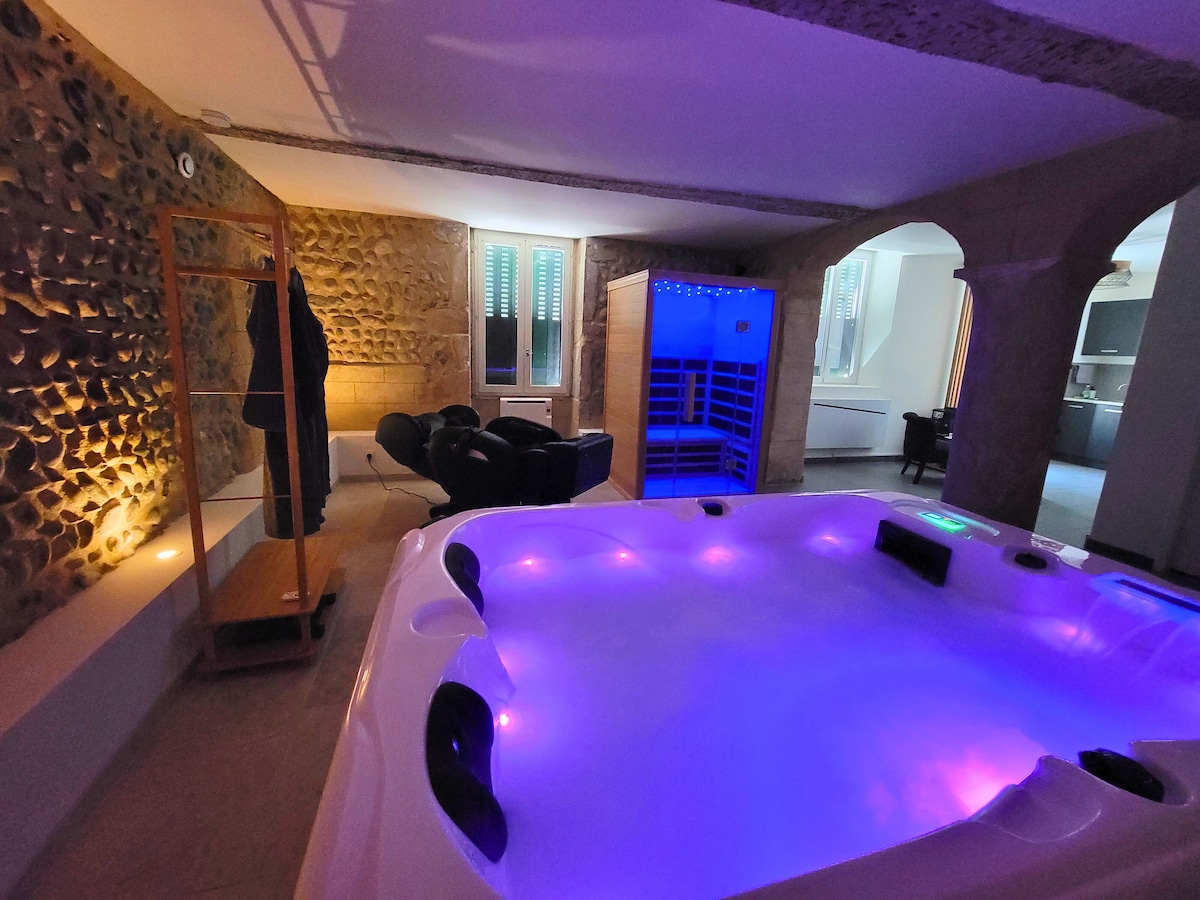 A spacious private jacuzzi is illuminated with subtle blue and purple lights, set against a backdrop of textured stone walls. Nearby, comfortable seating is arranged for relaxation, while doorway openings lead to a cozy kitchen area visible in the background.