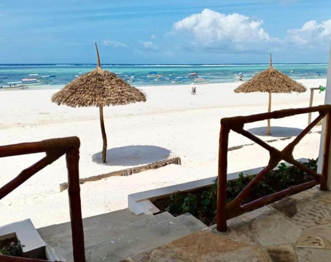 A view of the beach from the terrace showcases two thatched umbrellas on the sandy shoreline. The clear blue waters gently lap against the beach, with boats visible in the distance under a vibrant sky. The scene evokes a feeling of tranquility and openness.