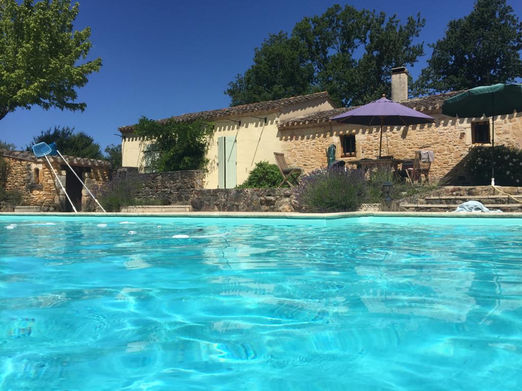 A clear blue pool reflects the sunny sky, framed by lush greenery. A stone property with bright yellow walls is visible in the background, featuring a shaded seating area beneath a purple umbrella. A tranquil setting invites relaxation and enjoyment of the outdoor space.