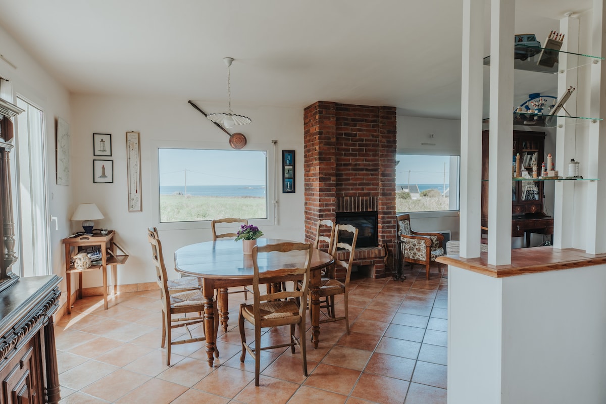 A dining area is shown with a wooden table surrounded by chairs, providing a view of the ocean through large windows. A brick fireplace adds warmth to the space, while terracotta tiles enhance the inviting atmosphere.