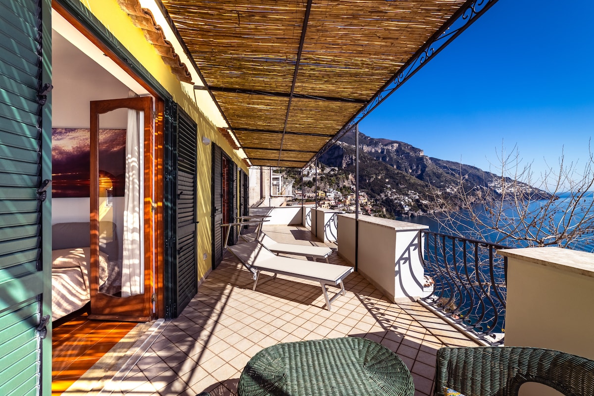 A sunlit terrace is presented with lounge chairs, offering expansive views of the coastline. The terrace's railing and nearby mountain landscape are clearly visible, along with an inviting seating area featuring wicker furniture. Adjustable awnings provide shade against the bright blue sky.
