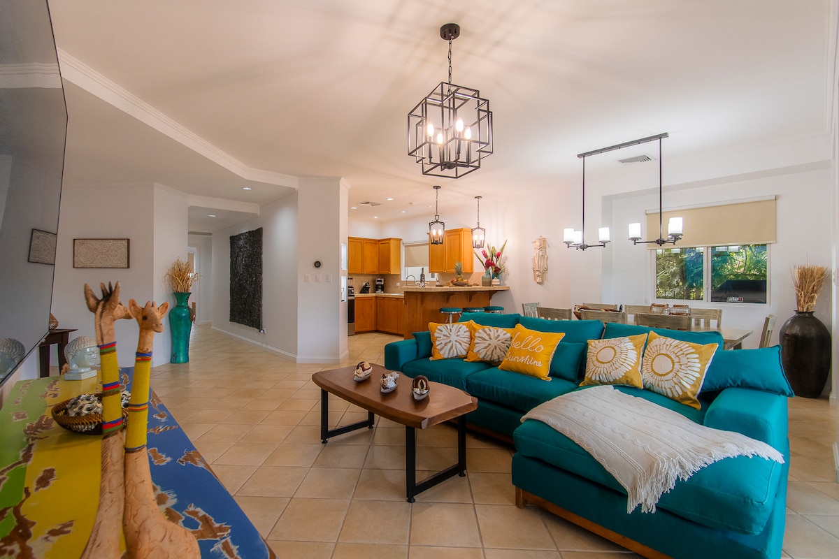 An open concept living area is highlighted by a spacious teal sectional sofa, adorned with colorful cushions. A wooden coffee table sits in front, complemented by decorative items. The kitchen area features warm wooden cabinetry and modern appliances, while ample natural light filters in through the nearby windows.