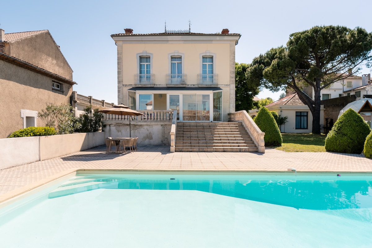 The elegant house showcases a spacious outdoor area featuring a large pool with clear blue water. Steps lead from the pool to the house, which has multiple large windows and a well-maintained garden. Surrounding greenery enhances the tranquil setting.