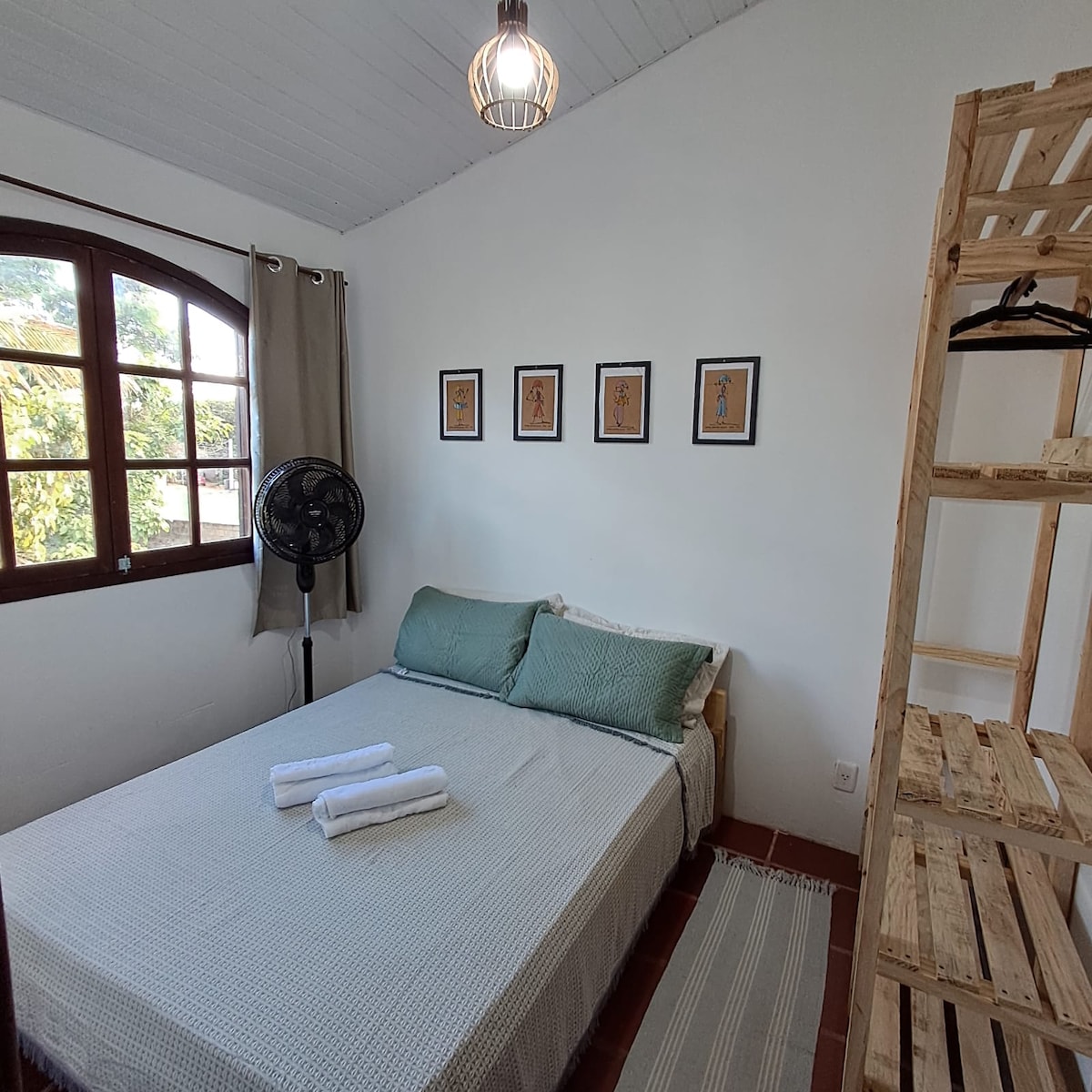 A bright bedroom is presented with a spacious bed adorned with crisp linens and folded towels. Natural light filters in through an arched window, complemented by decorative wall art. A standing fan and a wooden shelf with hangers contribute to the room's functional design.