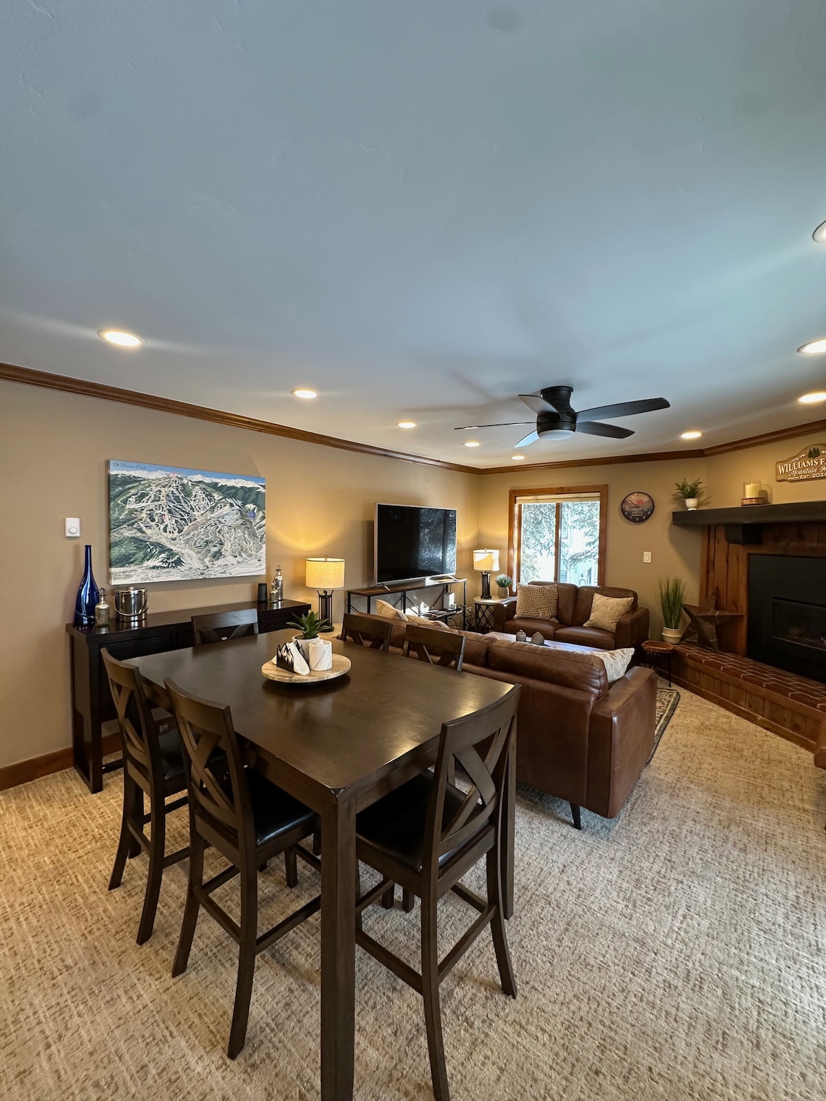 A cozy living area is featured with a comfortable seating arrangement surrounding a fireplace. A dining table with ample seating is positioned nearby. Natural light filters in through the windows, complemented by warm overhead lighting, creating a relaxing atmosphere.
