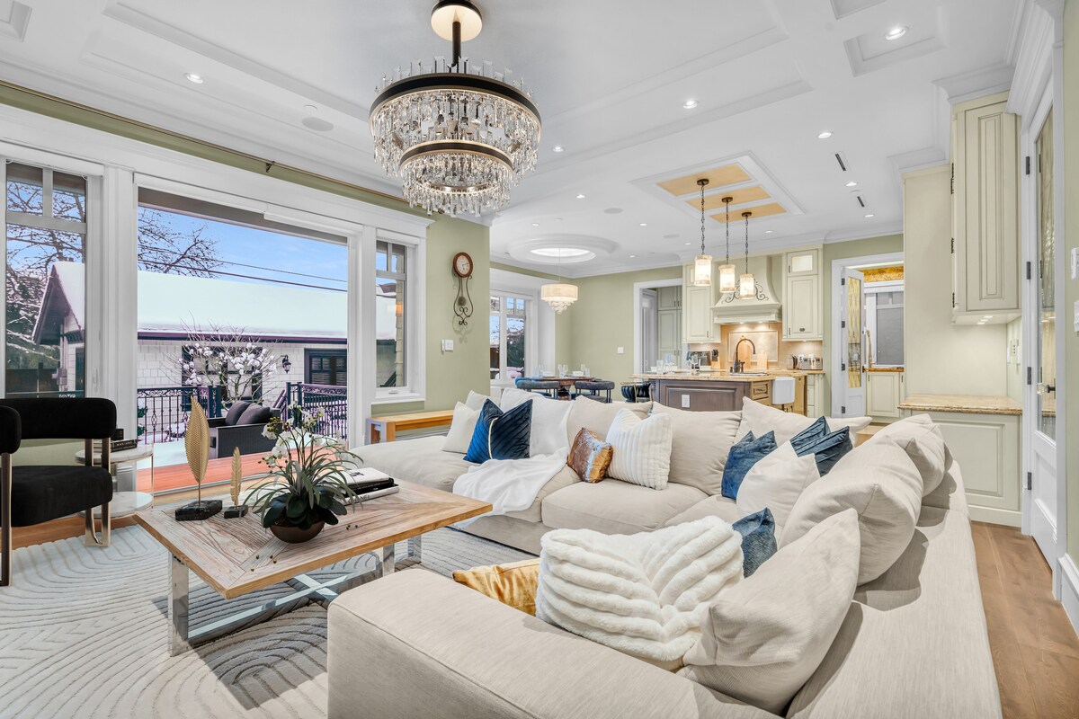 A spacious living room features a large sectional sofa in a light tone, arranged around a wooden coffee table. A chandelier hangs from the ceiling, and large windows provide natural light. In the background, a dining area is visible, showcasing a wooden dining table.