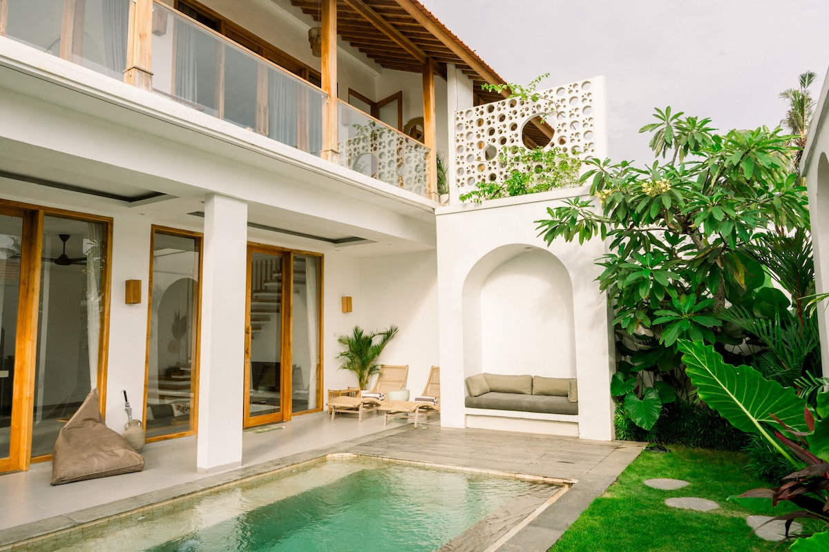 A private pool is surrounded by a well-manicured garden featuring lush greenery. The villa’s exterior showcases smooth white walls and warm wood accents, with expansive glass doors leading to the indoor spaces. A cozy seating area is present beside the pool.