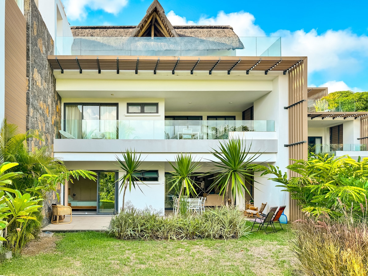 The exterior of a modern two-story apartment is depicted, featuring a spacious balcony with glass railings. Lush greenery surrounds the structure, including tropical plants and a vibrant lawn. Clear skies complement the contemporary architecture, suggesting an inviting atmosphere for relaxation.