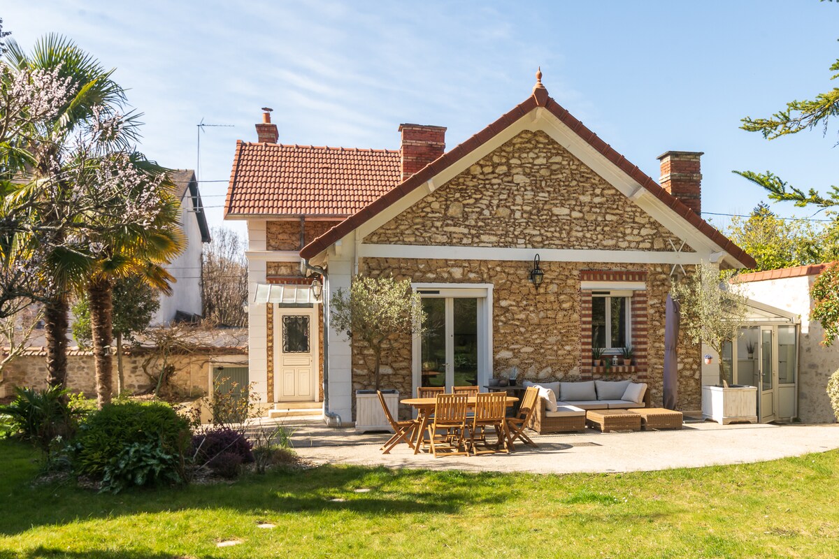 The exterior of the property is showcased, featuring a charming stone facade with a red-tiled roof. A spacious patio is highlighted, furnished with wooden dining furniture and a cozy seating area. Lush greenery and outdoor plants create a serene atmosphere in the garden.