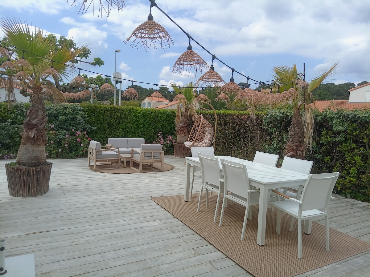An outdoor terrace features a spacious seating area with a beige couch and a circular table surrounded by six white chairs. Decorative lanterns hang overhead, while palm trees and lush greenery provide a natural backdrop.