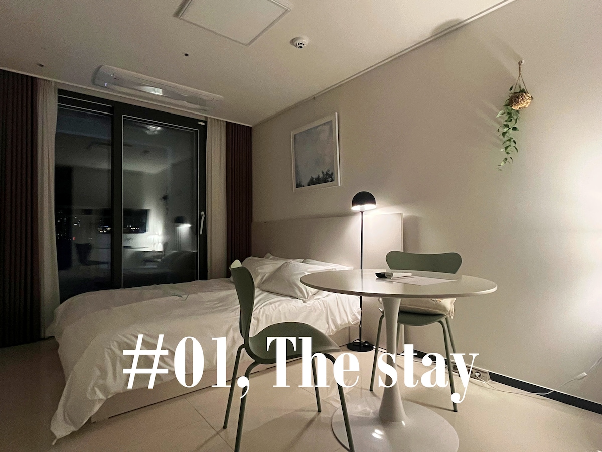 A studio room features a spacious bed dressed in soft bedding. A small round table is accompanied by two green chairs. A modern lamp provides warm lighting, and large windows reveal city views, allowing natural light to flow into the room.