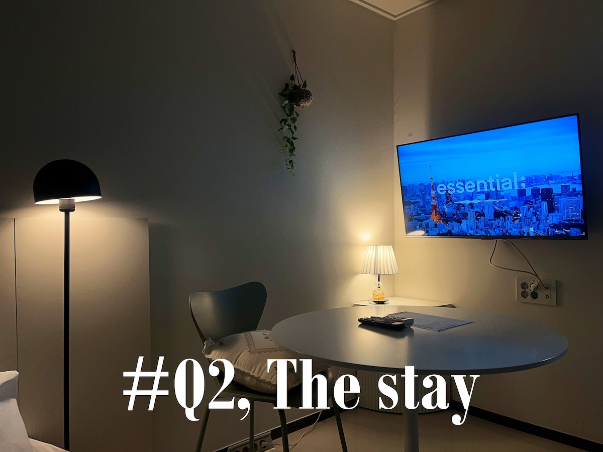 A modern studio room features a round dining table beside a comfortable chair. Soft lighting is provided by a floor lamp and a small table lamp. A wall-mounted television displays a serene urban view, creating a relaxing ambiance. Neutral tones contribute to the overall calm aesthetic.