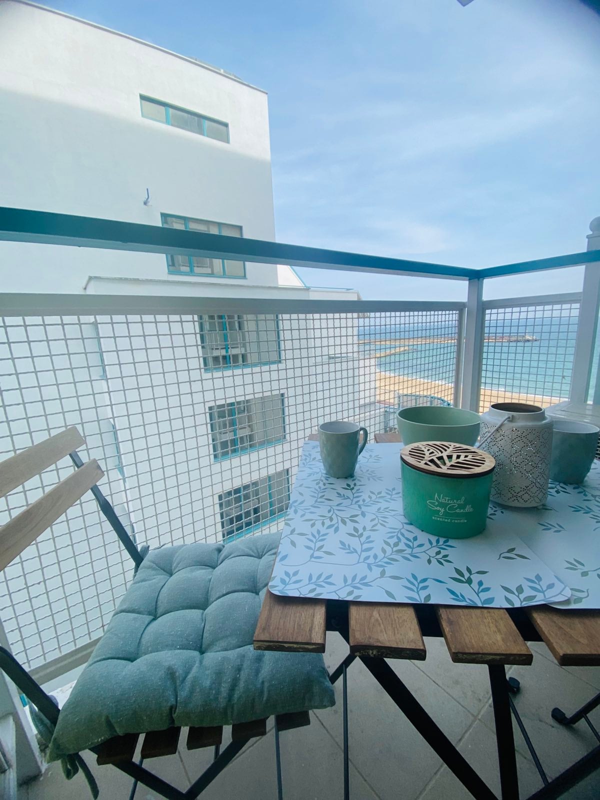 A small balcony features a wooden table and two chairs, surrounded by a protective railing. Green cushions are placed on the chairs. A decorative table setting includes a patterned tablecloth, a teal container, a candle holder, and two mugs. The ocean is visible in the background.