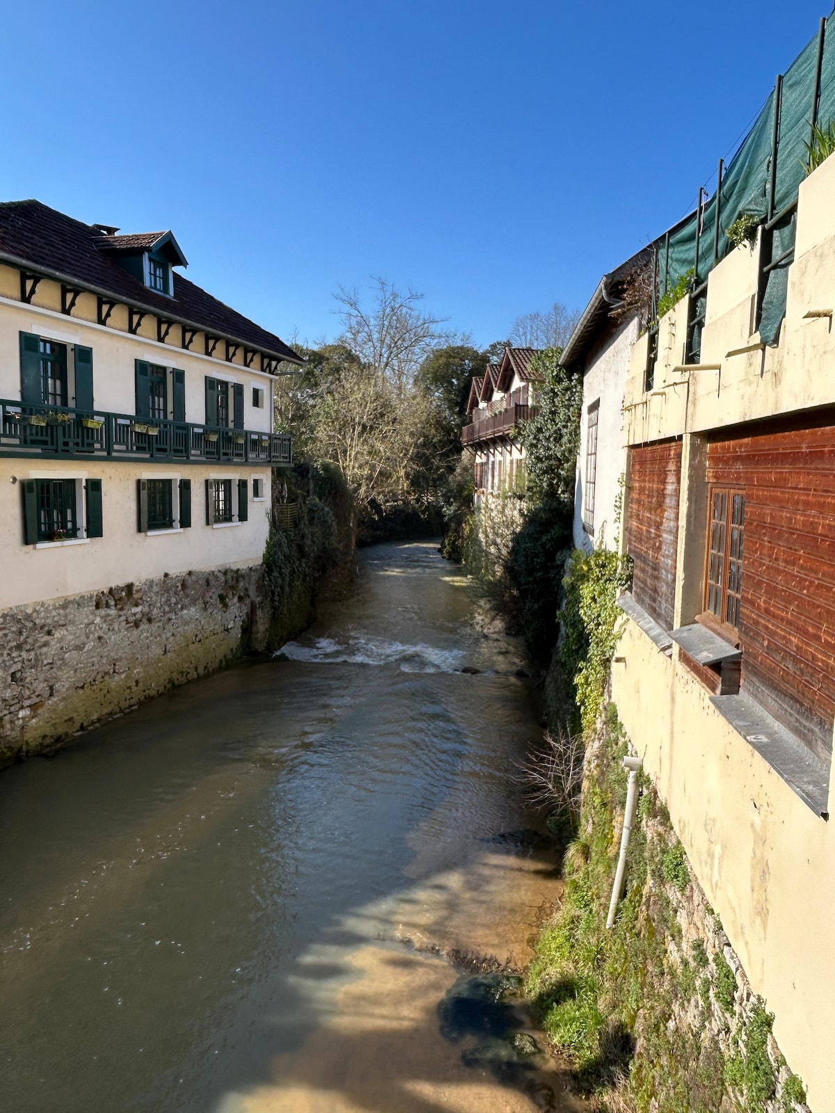 A serene view captures a narrow river flowing between two buildings. One structure displays charming wooden shutters and balconies, while the other features a large wooden window. Lush greenery lines the riverbank, enhancing the peaceful surroundings.