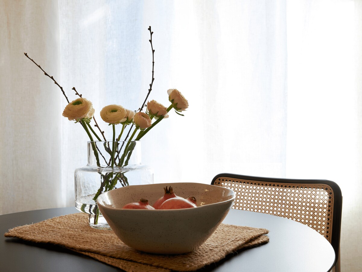 A dining table is set with a textured woven placemat featuring a bowl containing pomegranates. A clear glass vase holds delicate flowers, while soft light streams through sheer curtains, creating a calm and inviting ambiance.