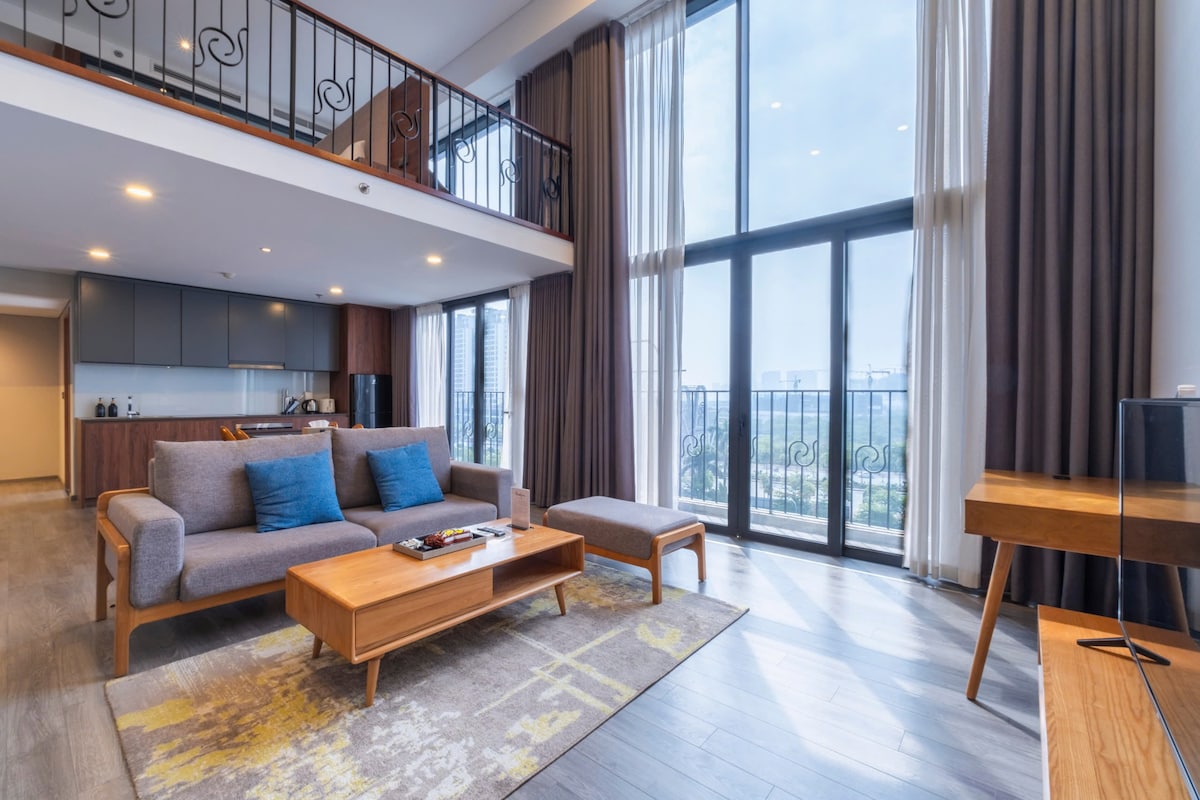The spacious living area features large floor-to-ceiling windows providing ample natural light and offering a view of West Lake. A modern couch with accent pillows faces a wooden coffee table, while a stylish rug adds warmth to the room. A well-equipped kitchen is visible in the background.