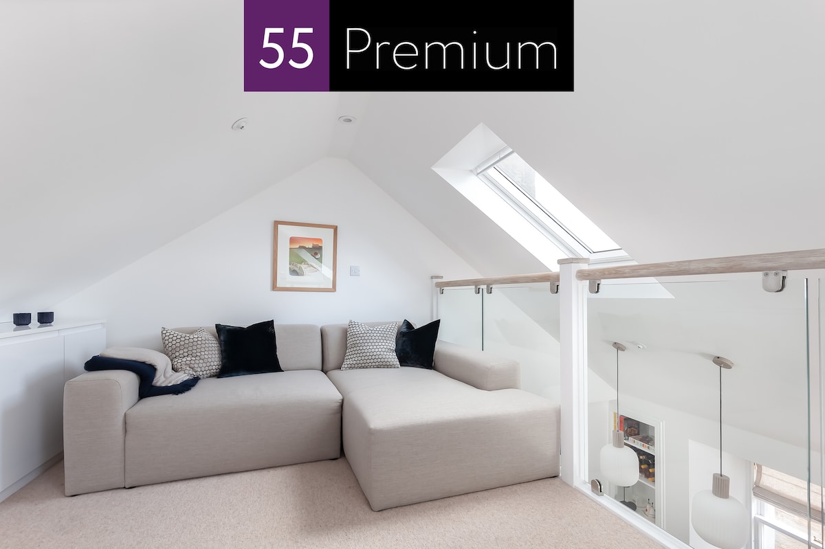 A light-filled mezzanine lounge area is highlighted by a large, modular sofa in neutral tones. Natural light floods in through a skylight, illuminating the soft carpeted floor. Minimalistic decor and sleek glass railings contribute to the contemporary design of the space.