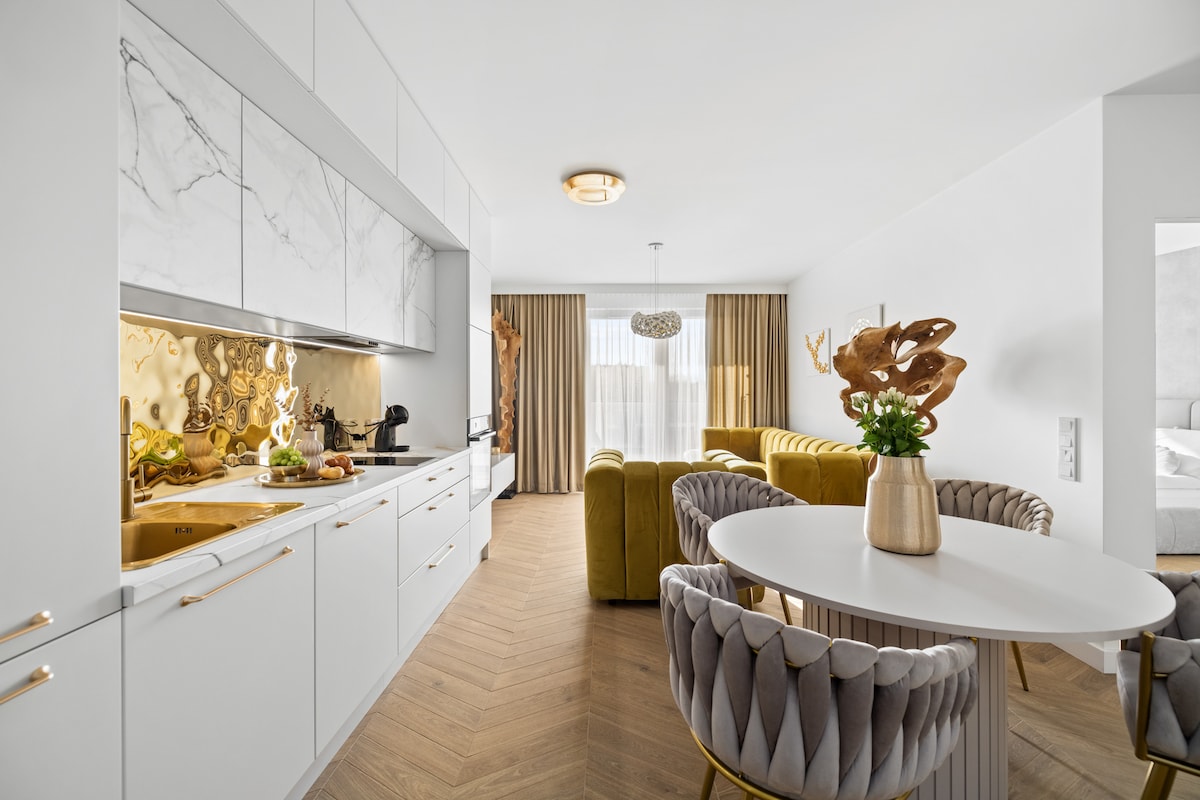 A modern kitchen is presented featuring white cabinetry with marble accents. A round dining table with plush, upholstered chairs is positioned near a window. The open-plan layout reveals a cozy living area with a yellow sofa and decorative elements that enhance the overall contemporary design.