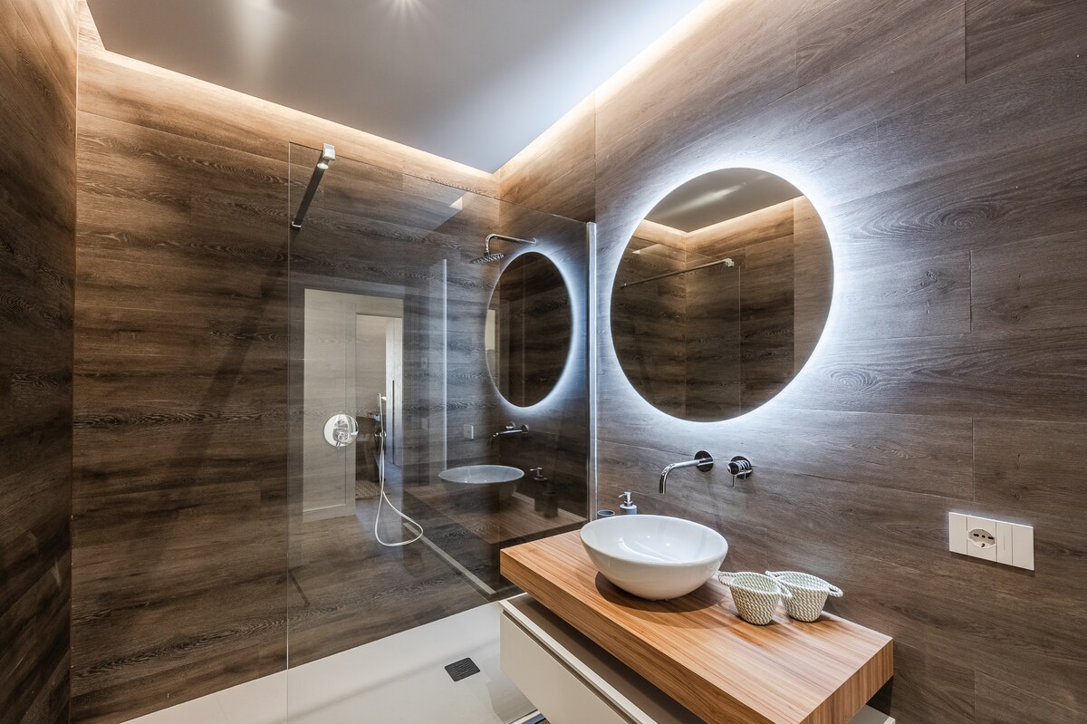 An elegant bathroom features a spacious walk-in shower enclosed by glass walls. Two round mirrors are illuminated with soft lighting above a modern vanity with a wooden countertop and a bowl sink. Natural wooden textures create a warm ambiance.