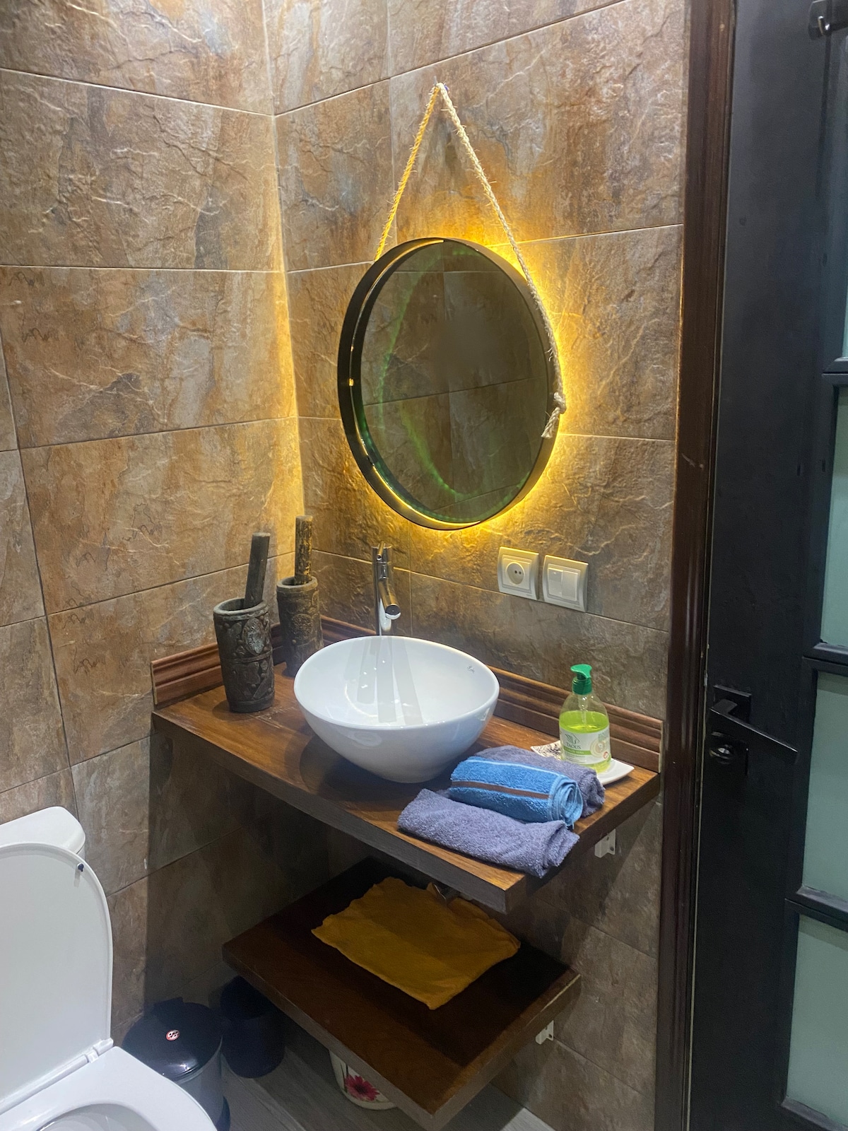 A modern bathroom setup features a circular mirror illuminated by soft lighting. A sleek white sink is positioned on a wooden shelf, accompanied by two decorative vases. Clean towels are neatly arranged, and a liquid soap dispenser is placed beside the sink.