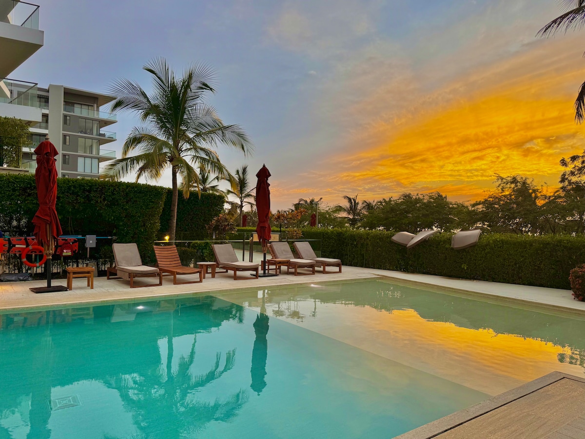 A serene pool area is surrounded by lush greenery, with lounge chairs positioned neatly along the edge. Umbrellas are placed strategically for shade, and a soft sunset casts warm colors across the water, reflecting the vibrant sky.