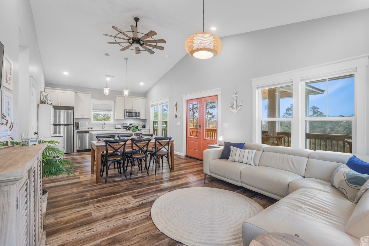 An inviting open-concept living area showcases a spacious sectional sofa, a round area rug, and a dining table set for six. Large windows allow natural light to fill the space, highlighting the modern kitchen with stainless steel appliances. A front door painted in a warm coral adds a touch of charm.