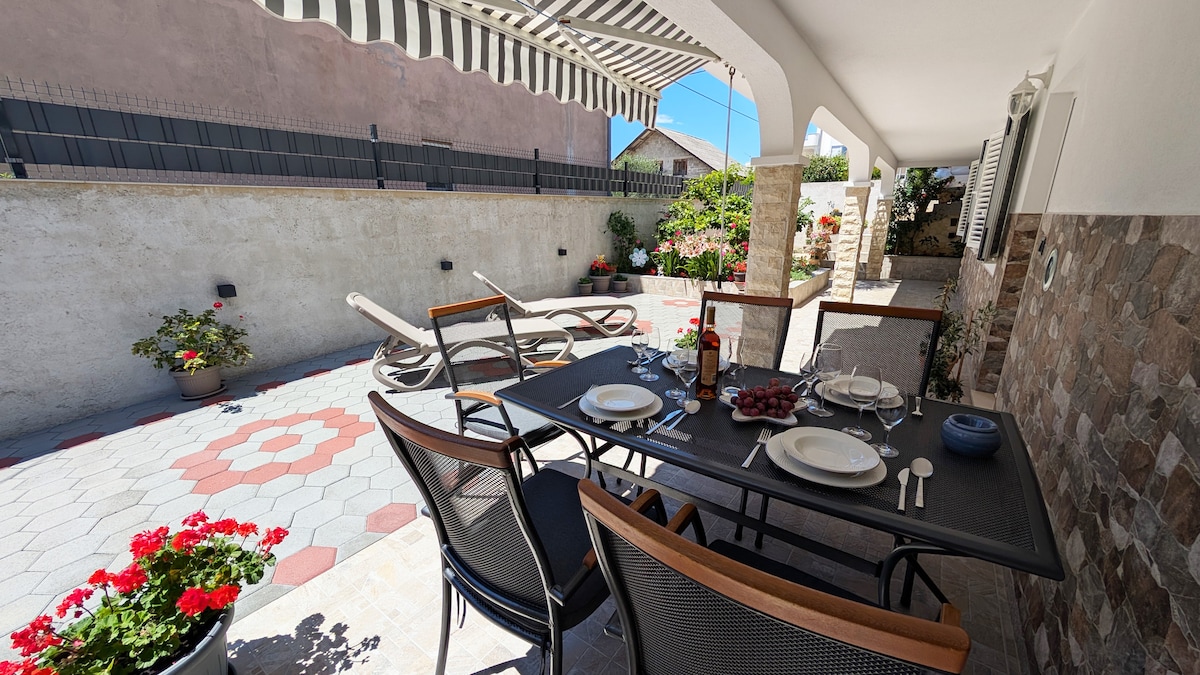 An outdoor terrace features a dining table set for six, accompanied by comfortable chairs. Sun loungers are positioned nearby, providing space for relaxation. Lush greenery and colorful flowers add charm to the area, while shaded seating offers a comfortable spot to enjoy meals or drinks.