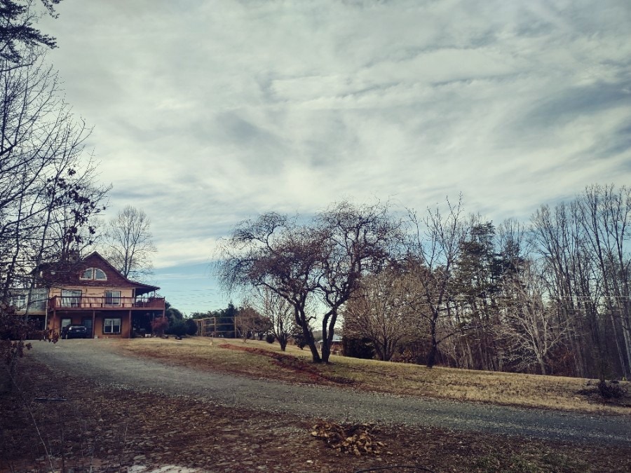 A scenic view showcases a spacious home set against a tranquil sky, with wispy clouds above. A gravel driveway leads to the house, bordered by a leafless tree and open green areas, creating a peaceful outdoor setting.