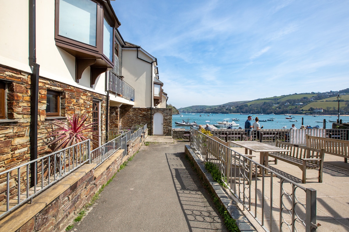 A path lined with low stone walls leads towards the water's edge, showcasing the estuary with boats anchored in the distance. Sunlight illuminates the area, while mixed materials of stone and wood are visible on the nearby buildings.