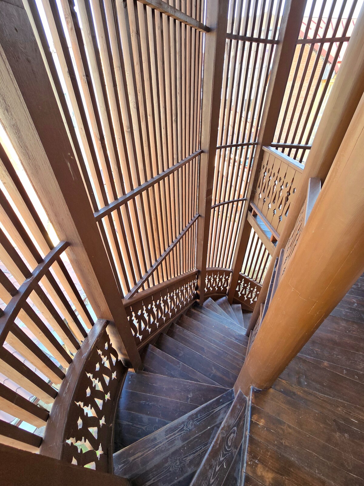 A spiral staircase with wooden steps winds gracefully downward. The staircase features intricately designed wooden slats that create a play of light and shadow, enhancing the architectural detail of the space. The warm tones of the wood add a sense of warmth and character to the environment.