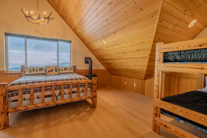 Спална соба на горниот кат со брачен кревет (широк 150-179 см) и двокреветна соба. Заоблените тавани, рустикалниот мебел од трупци и големите прозорци носат природна светлина и поглед кон планината - совршени за мирни ноќи и опуштени утра.