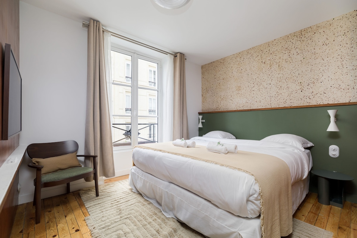 A spacious sleeping area features a king-size bed with a light-colored duvet and neatly arranged towels. Soft light enters through large windows, covered by light curtains. A wooden chair is positioned nearby, and the room is accented by textured walls and simple, elegant decor.